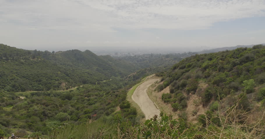 Cinematic daytime 4K 10-bit DSLR footage captured from a Griffith Park hilltop looking down toward Hollywood. The shot features equestrians on horseback slowly descending a canyon fire road.