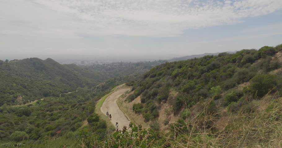 Cinematic daytime 4K 10-bit DSLR footage captured from a Griffith Park hilltop looking down toward Hollywood. The shot features equestrians on horseback slowly descending a canyon fire road.