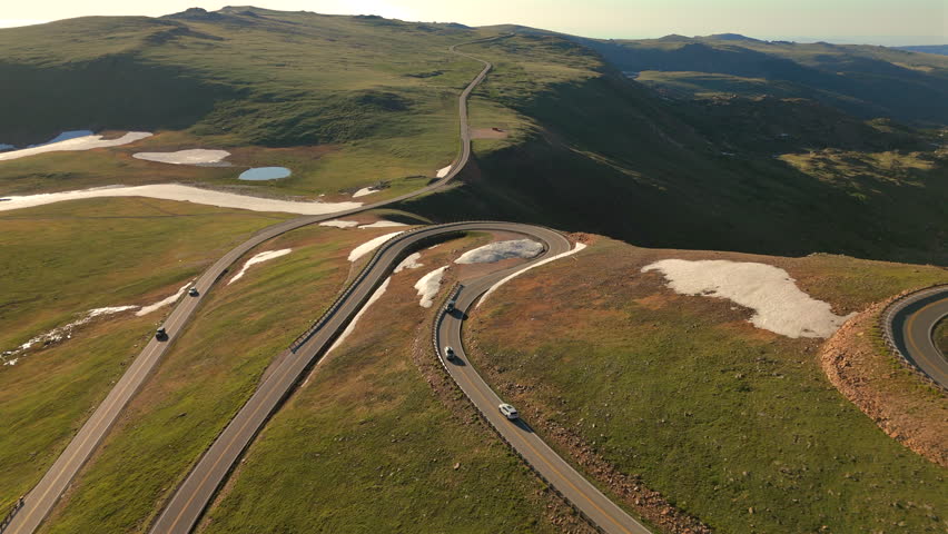 Beartooth Highway in Montana winds through alpine terrain with dramatic hairpin curves. Cars travel along the scenic mountain pass with patches of snow and green meadows visible from above.