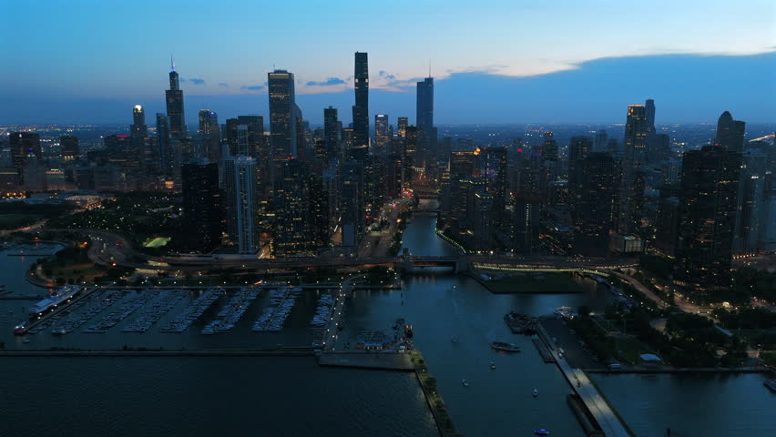 Stunning aerial footage of Chicago downtown skyline at twilight with illuminated skyscrapers along the Chicago River. Navy Pier ferris wheel glows as boats dock in the marina harbor below.