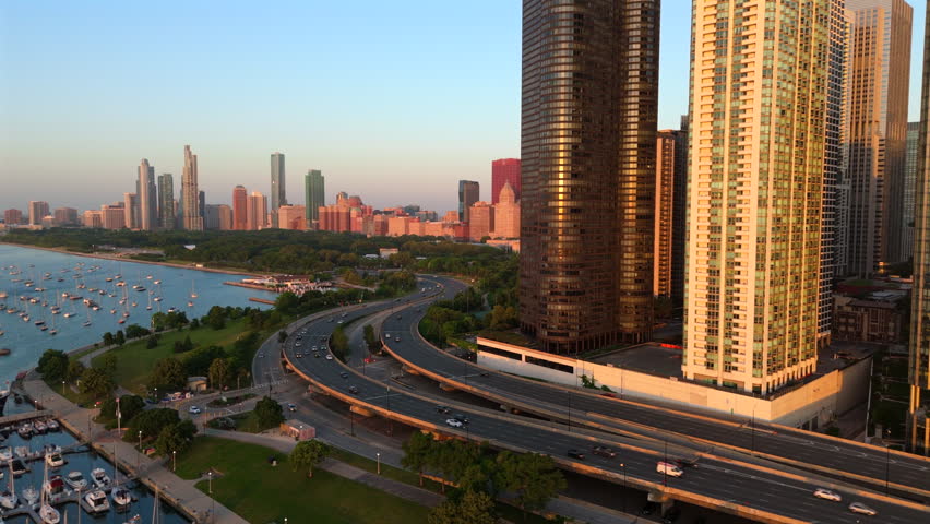 Aerial view of Chicago skyline during golden hour featuring Lake Shore Drive highway curving along Lake Michigan. Modern skyscrapers reflect warm sunlight while boats dock at marina below.