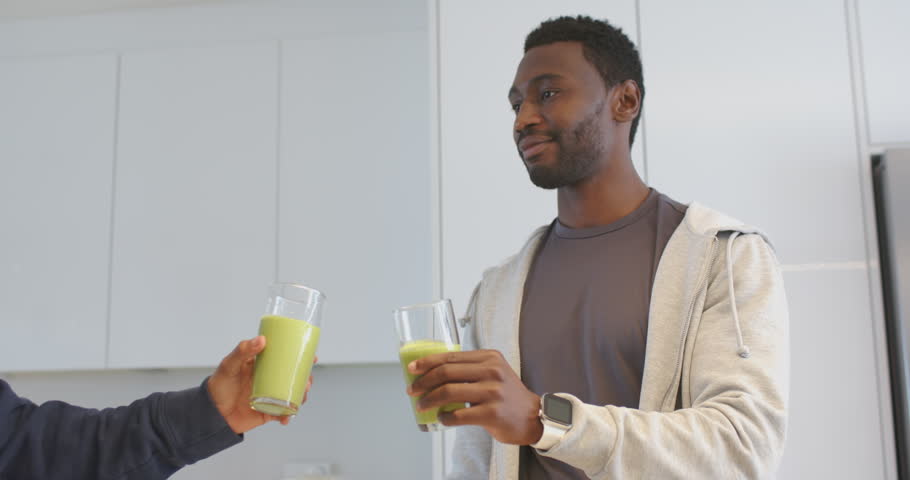 Diverse male friends tapping glasses in kitchen and sipping green smoothies for healthy moment. Health, wellness, friendship, casual, modern, lifestyle, organic