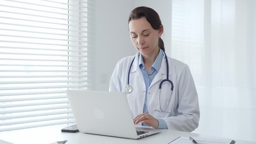 Female doctor physician working on laptop, wearing white lab coat and stethoscope, seated at desk in bright, sunlit medical workspace. Medicine and health care concept