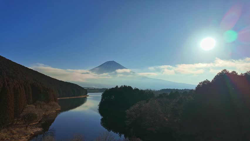 A breathtaking sunrise view of Mount Fuji over Lake Tanuki. Features a brilliant sun flare, soft morning mist, and the silhouettes of surrounding evergreen forests in Japan.
