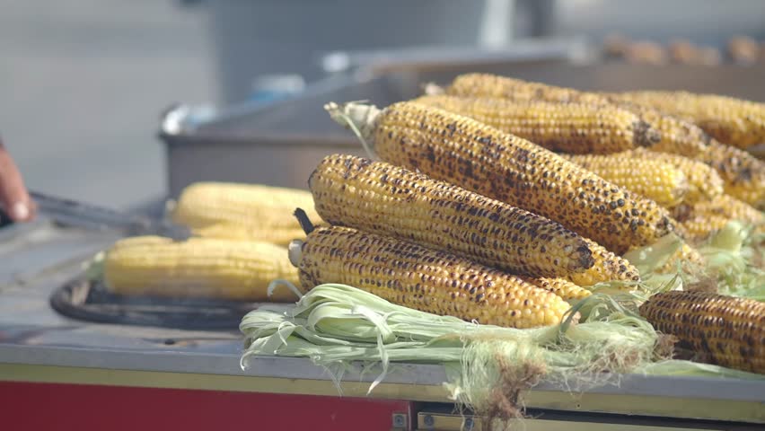 Close up shot of golden corn cobs roasting on charcoal grill vendor using tongs to flip vegetables at festival stall.