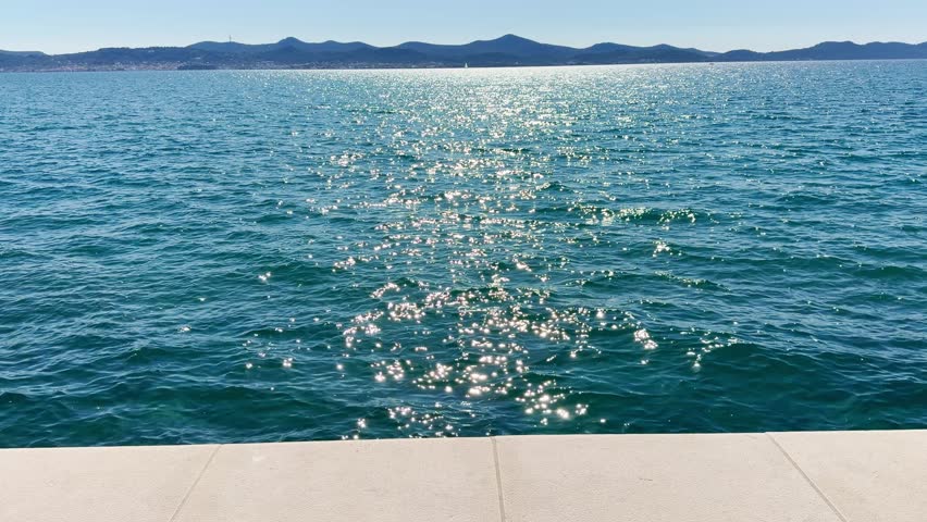 Slow‑motion shot of the shimmering turquoise Adriatic in Zadar, with a thin white stone promenade edge for contrast and distant Ugljan island on the bright horizon.