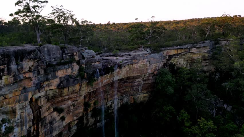 Aerial footage of waterfalls cascading over rocky cliffs in rural New South Wales, Australia, surrounded by lush greenery, captured in summer.