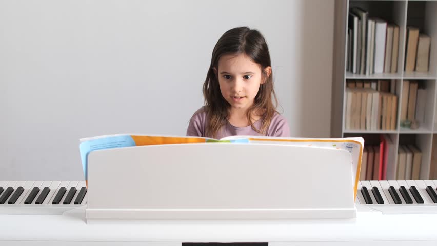 Curious young girl concentrating and reading sheet music from an open book on a digital piano stand, enhancing her music education and skill development during a lesson