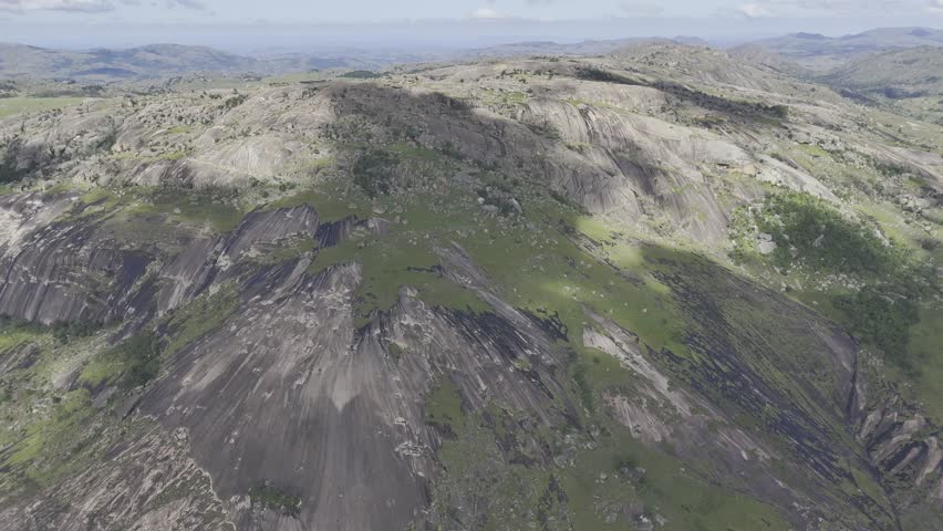 Drone orbits to the right in ultra wide shot of the western face of the rocky summit under cloud cover on a sunny day at Sibebe Rock near Mbabane, Eswatini
