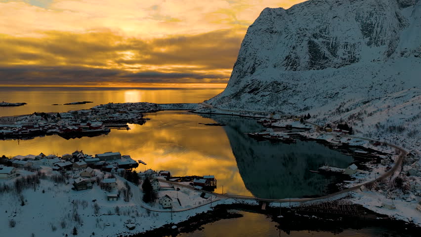 Breathtaking drone shot of Reine village in Lofoten, Norway, featuring snowcapped mountains reflecting in calm arctic waters during a vibrant sunset.