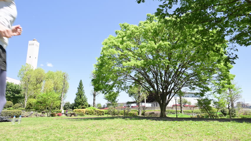 A wide-angle, full-body video of a woman engaging in aerobic exercise and running in a park with fresh greenery under a blue sky, capturing a healthy lifestyle through sports photography—a spring summer image featuring short sleeves