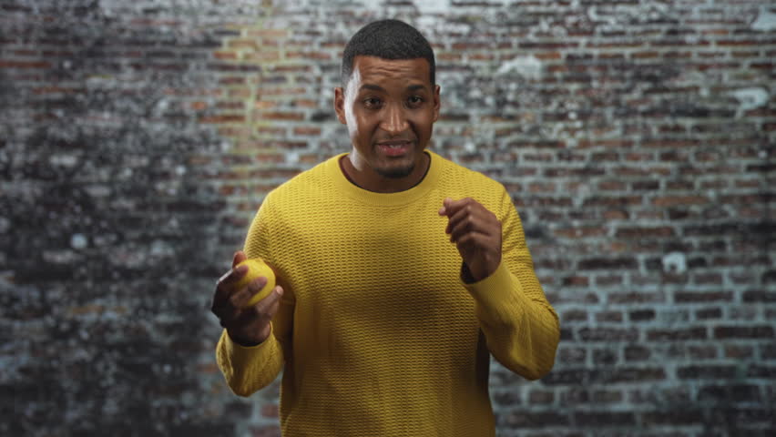Black man holding a lemon with hands and gesturing near face in a studio with brick wall; curiosity freshness simplicity.