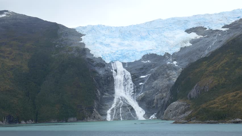 A large waterfall flows down from a glacier in Patagonia. The glacier sits high on a mountain. The lake below reflects the surrounding nature. Clouds are present in the sky.
