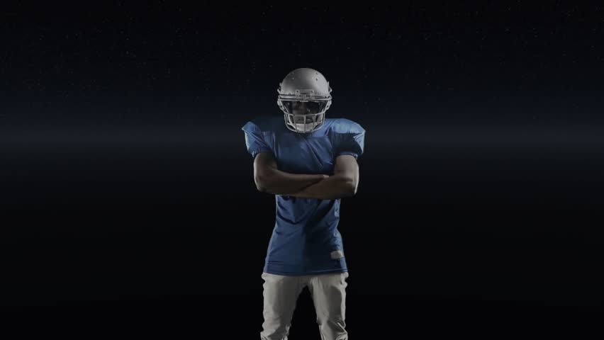 Male football player in helmet uncrossing arms and pointing, revealing sports HUD with bar charts. Holographic, futuristic, athlete, studio, headgear, pads, blue