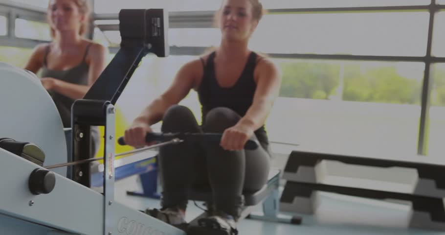2 women rowing in gym camera pan left showing flywheel animation tracking workout near purple ball. Ergometer, duo, cardio, fitness, studio, sunlit, rhythm
