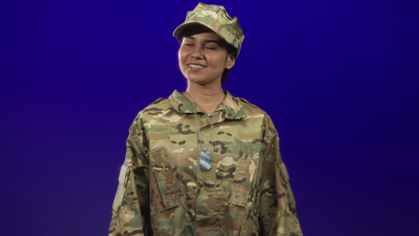 Woman soldier in camouflage uniform smiles and gives thumbs up, dog tags visible, slight head turn in studio; pride duty.