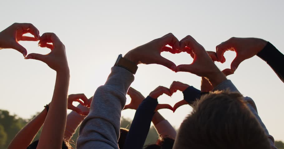 Group of unrecognizable young people doing heart gestures against bright sunny sky background. Friends holding up hands making heart shapes in sunlight. Love and friendship concept