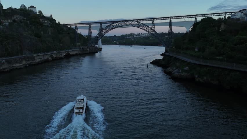 Aerial view of a boat sailing under the D. Maria Pia bridge across dark waters, contrasting with the lights of the city at dusk, Porto, Portugal.