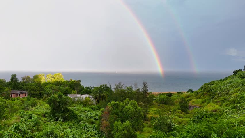 Stunning Double Rainbow Over Ocean and Greenery, Rare sight of a double rainbow stretching over lush green hills after rain.