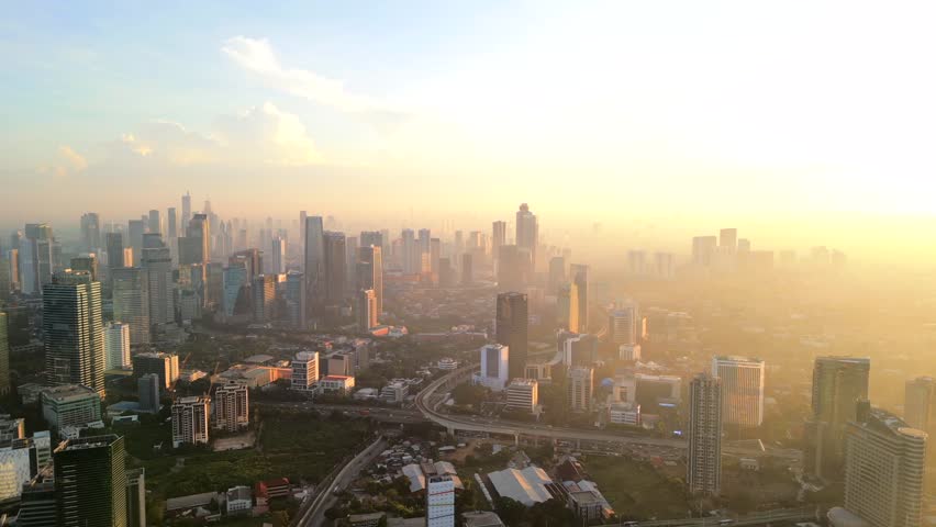 Aerial view of Jakarta skyline at sunrise with warm golden light and morning haze. Cinematic cityscape showing urban density, modern buildings and early day atmosphere.