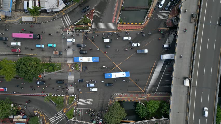 Aerial top-down view of busy X-shaped intersection in Jakarta, Indonesia. Urban traffic with cars, motorbikes, buses, crosswalks, and elevated highway infrastructure.
