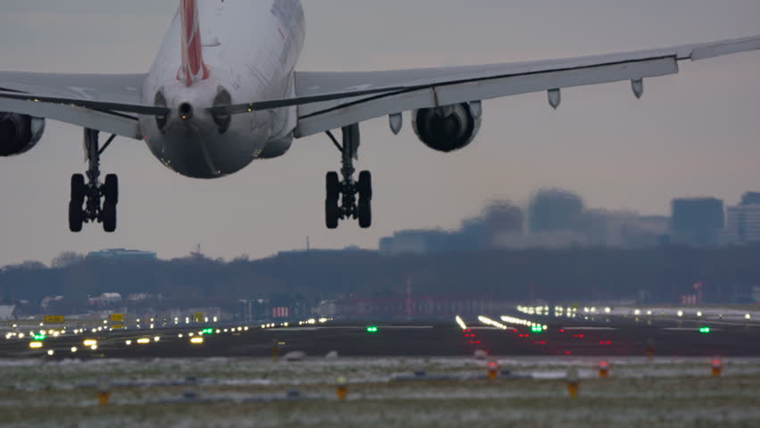 A large passenger airplane lands on an airport runway. The video shows a rear view of the aircraft descending, touching down, and rolling along the illuminated tarmac during a winter sunset