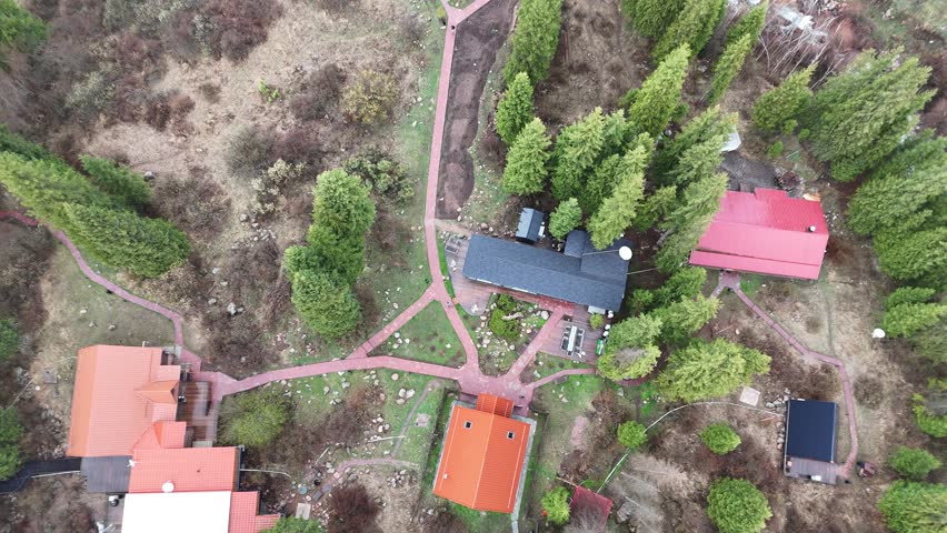 Quick aerial drone footage descending over a private property with a garden and surrounding evergreen trees. The video offers a direct top-down view, highlighting red-roofed houses, paved walkways, outbuildings, and landscaping details. The fast descent provides a fresh perspective over residential buildings and green outdoor spaces—ideal for use in real estate, architecture, property promotion, drone technology, and nature-meets-urban concepts.

