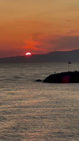 Sunset over sea with sun setting behind distant island silhouette, dark water and warm orange sky, gentle motion creating changing coastal view with foreground land and small cross visible on it, evening seascape, Cyclades Greece
