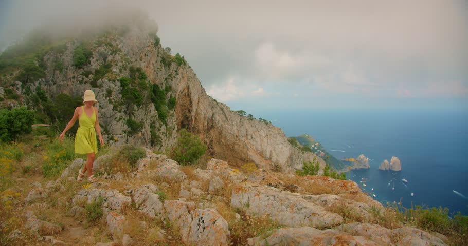 A woman walks over rocky terrain on a high cliff on Monte Solaro above Faraglioni sea stacks in Capri Italy