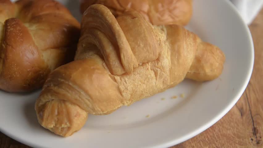 Croissant cut in half to be spread with lots of strawberry jam on a white plate on a wooden table, macro or close-up view from a horizontal angle