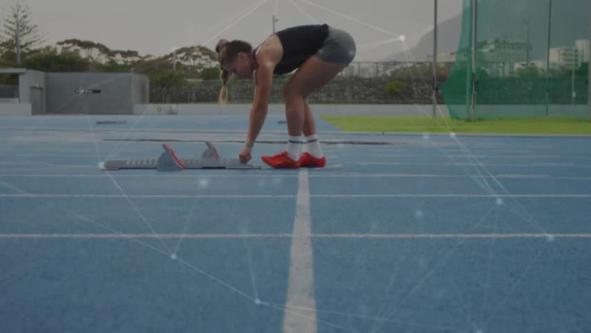 Sprinter adjusting blocks on blue track, preparing for sprint while particle overlay tracing motion. Runner, outdoors, training, focus, spikes, watch, athletics