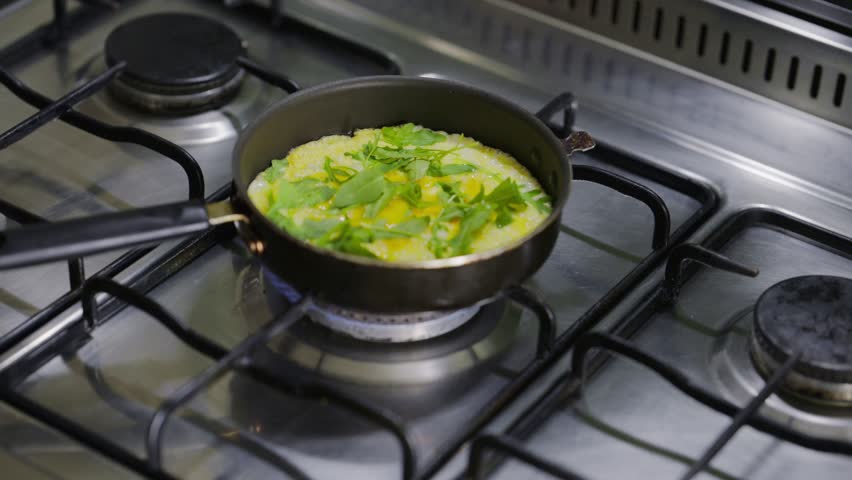 Close up view of a person using a yellow spatula to fold a delicious, fluffy omelet filled with fresh arugula while it cooks in a small frying pan over a gas burner in a kitchen