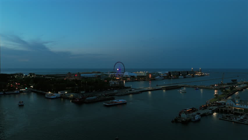 Aerial drone footage captures Navy Pier waterfront at twilight with illuminated ferris wheel glowing purple against darkening blue sky, boats drifting through calm harbor waters reflecting city.