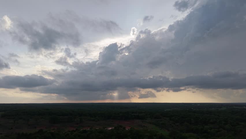 Drone aerial push forward over forest and pond toward storm with rain shafts at sunset in Oklahoma.