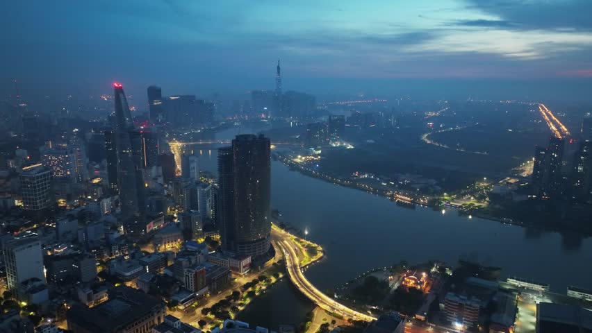 High angle drone shot capturing curving river and bright road network with modern skyline fading into atmospheric haze at dusk in Saigon.