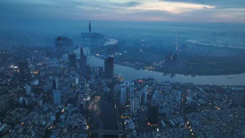 Expansive drone view over Saigon with river bending through dense buildings and soft glowing city lights beneath early night sky.