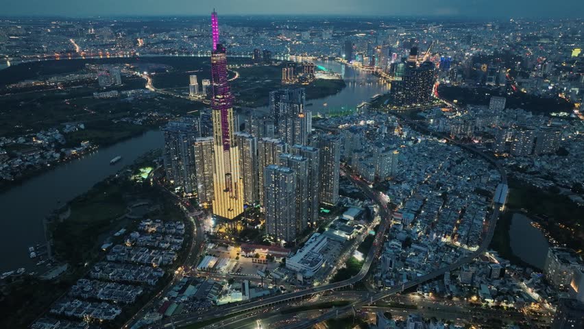 High altitude drone shot highlighting clusters of high-rise buildings surrounded by dense urban blocks, highway intersections, and glowing riverside skyline at evening in Saigon.