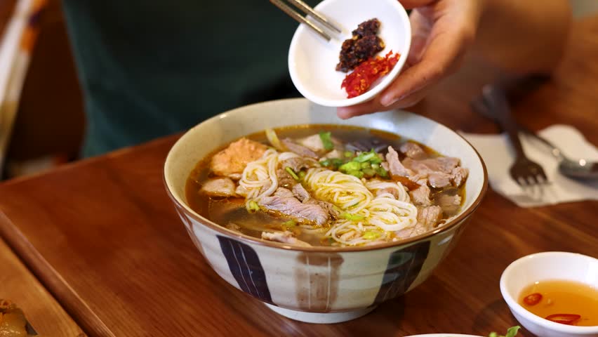 A person adds spicy chili oil to Vietnamese bun cha noodles in a restaurant setting