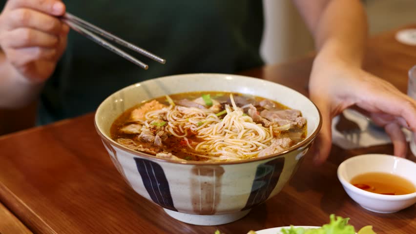 Close-up shot of hands pouring fish sauce into a ceramic bowl of traditional Vietnamese noodles