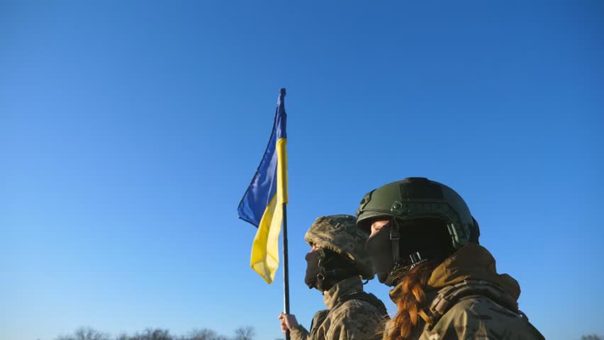 Military couple in camouflage standing on a hilltop holding hands, raising a flag, looking at the sky in a cinematic sunrise scene.
