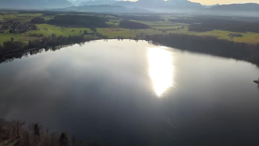 Calm winter view of a lake from above, surrounded by rural land, trees and distant hills.
