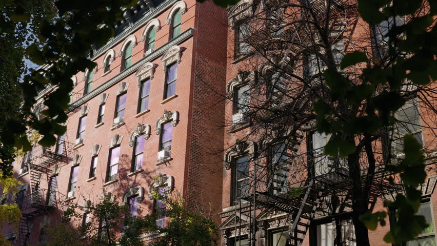 Historic red brick apartment houses with ornate windows and fire escapes framed by trees, classic New York City neighborhood street architecture.