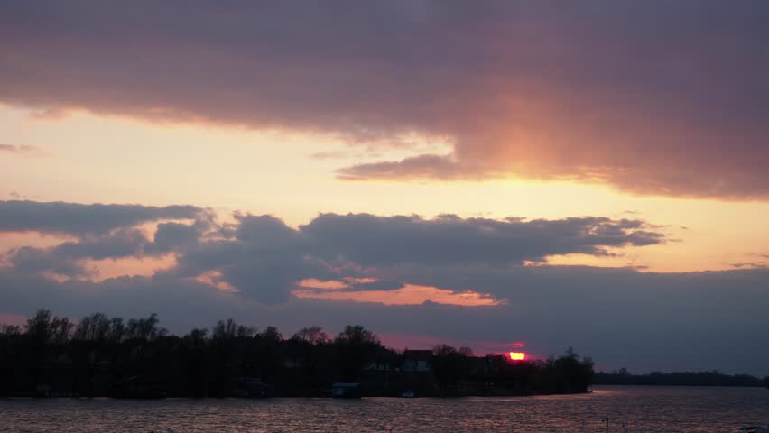 The sun sets over the lake as clouds fill the sky. Colors change from orange to purple. The view shows the lake shore and trees just before nightfall