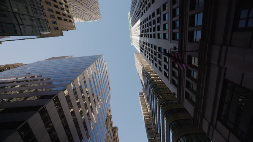 Low-angle street-level view looking up between towering modern skyscrapers in Manhattan, New York City, USA. Glass facades and geometric architecture frame the blue sky above.