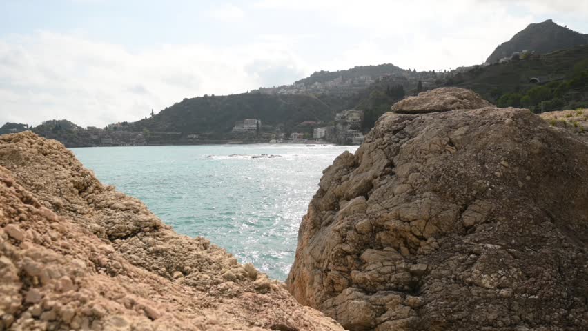 View of a rugged coastline with large rocks in the foreground and a town nestled on a hillside overlooking the sparkling blue sea