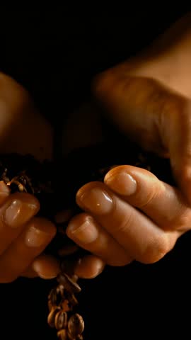 Vertical video: Woman cupping coffee beans on black, tilting fingers releasing beans to show coffee. Palms, roasted, cascade, closeup, macro, lowkey, vertical video