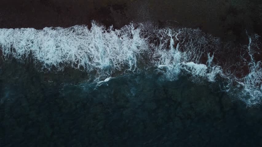 A dramatic top-down aerial view of dark blue ocean waves crashing onto a rocky shoreline with white foam.