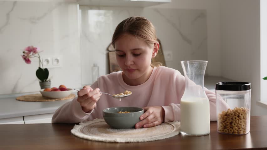 Young woman eats breakfast cereal from a grey bowl in a bright kitchen. Milk container sits on counter.