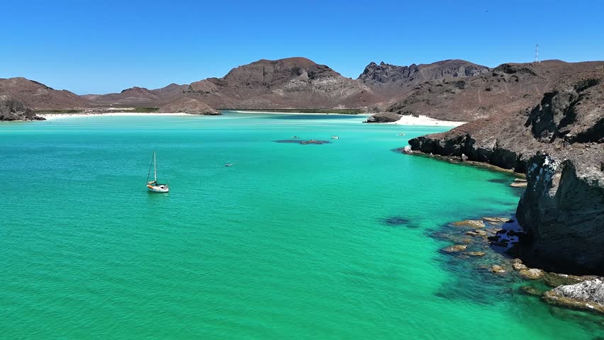 4K drone following a rocky cliff at Playa Balandra in La Paz toward sailboats and a small rocky island as birds lift off over calm water.