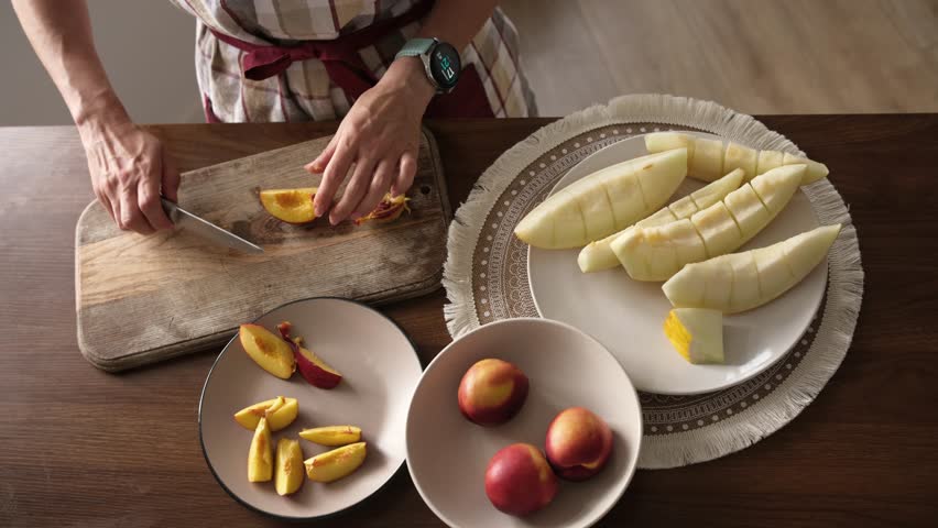 Girl slicing peaches on wooden board. Peeled apples and whole peaches sit on plates on kitchen table.